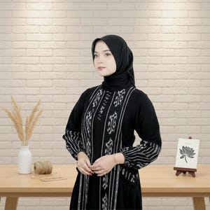 Puede incluir: Vestido negro con detalles de patrones verticales blancos. El vestido tiene mangas largas con puños a rayas. Un hiyab negro cubre la cabeza. Una pequeña tarjeta con una ilustración de una flor está en un caballete de madera.