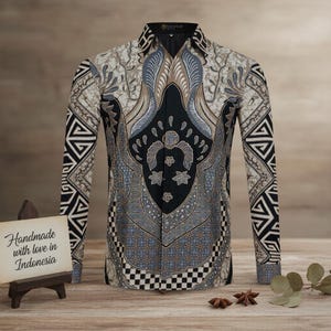 Op de afbeelding: Een overhemd met lange mouwen en een gedetailleerd batikpatroon in zwart, grijs en beige. Het overhemd heeft een geometrisch ontwerp op de mouwen en een centraal motief met bloemen en abstracte elementen. De tekst "Handmade with love in Indonesia" is zichtbaar.