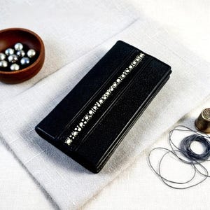 May include: A black rectangular case with a textured surface and a decorative white dotted stripe. The case is lying on a white cloth, with a small wooden bowl of silver beads and sewing notions in the background.