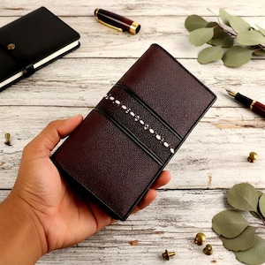 Könnte beinhalten: Eine dunkelbraune Ledergeldbörse mit strukturierter Oberfläche, zwei schwarzen Streifen und einem zentralen weißen Punktmuster. Die Geldbörse wird in einer Hand gehalten, im Hintergrund ein schwarzes Notizbuch und ein Stift.