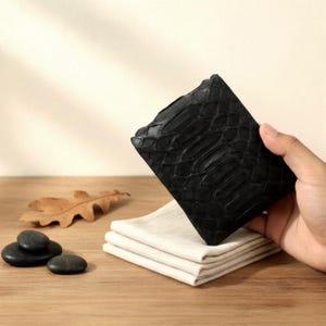May include: A black leather wallet with a textured, scale-like pattern. The wallet is held over a stack of folded white cloths. A few black stones and a dried leaf are on the wooden surface.