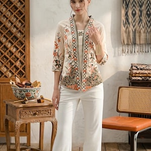 May include: A woman in a cream-coloured floral patterned jacket and matching top, with white trousers. The jacket has orange and green floral designs. The outfit is part of a fashion collection.