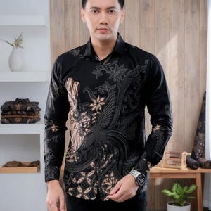 May include: Black long-sleeve button-down shirt with a detailed batik pattern in shades of brown and gray. The shirt features a classic collar and cuffs. The design includes floral and bird motifs. The shirt is paired with black pants.