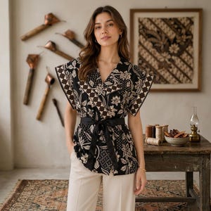 May include: A black and beige floral patterned short-sleeve blouse with a V-neckline and a black fabric waist tie. The garment features a wrap-style front with wide sleeves and a repeating botanical print, paired with light beige trousers.