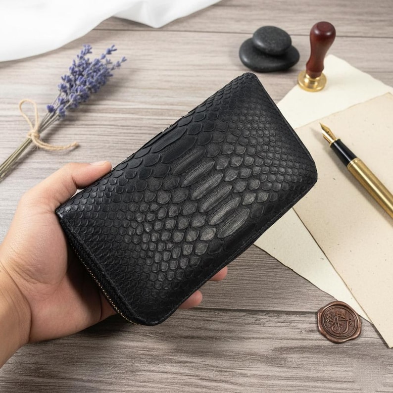 Puede incluir: Cartera de cuero negro con un dise&ntilde;o de piel de serpiente, sostenida por una persona. La cartera tiene un cierre de cremallera. Otros elementos en la superficie de madera incluyen un bol&iacute;grafo, un sello de cera y lavanda.