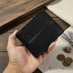 Puede incluir: Una cartera de cuero negro texturizado, de forma rectangular, con un detalle de doble línea. La cartera se sostiene en una mano, mostrando su diseño y artesanía. La cartera parece nueva y sin usar.