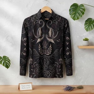 May include: A black long-sleeved shirt with a detailed batik pattern in shades of brown and cream. The shirt is hanging on a wooden hanger. The text on the tag reads "Handcrafted Batik Shirt."