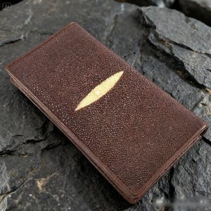 May include: A brown leather wallet with a textured surface and a light-colored, oval-shaped accent. The wallet is rectangular and appears to be closed. The background is a dark, textured surface.
