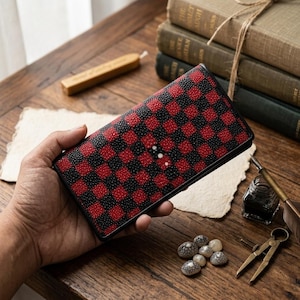 Könnte beinhalten: Eine schwarze und rote karierte Geldbörse. Die Geldbörse wird in einer Hand gehalten und hat einen schwarzen Rand. Die Geldbörse hat drei kleine weiße Punkte auf der Vorderseite. Die Geldbörse liegt auf einer Holzoberfläche.