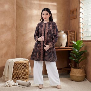 May include: A long-sleeved, open-front jacket with a brown and black batik pattern. The jacket is paired with white wide-leg pants. The outfit is worn with beige shoes. The background features a woven basket, a plant, and a wooden table.