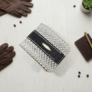 Könnte beinhalten: Eine rechteckige Brieftasche mit einem weiß-schwarzen Muster, mit einem schwarzen Streifen in der Mitte. Die Brieftasche ist umgeben von braunen Lederhandschuhen, einem kleinen Notizbuch, einem Stift und einer Topf-Sukkulente. Kaffeebohnen sind verstreut.