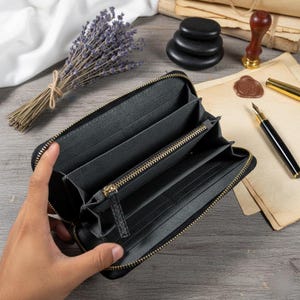 Puede incluir: Cartera negra abierta con m&uacute;ltiples compartimentos y cremallera dorada. La cartera est&aacute; hecha de un material oscuro. El fondo incluye un ramo de lavanda, piedras apiladas y art&iacute;culos de papeler&iacute;a.