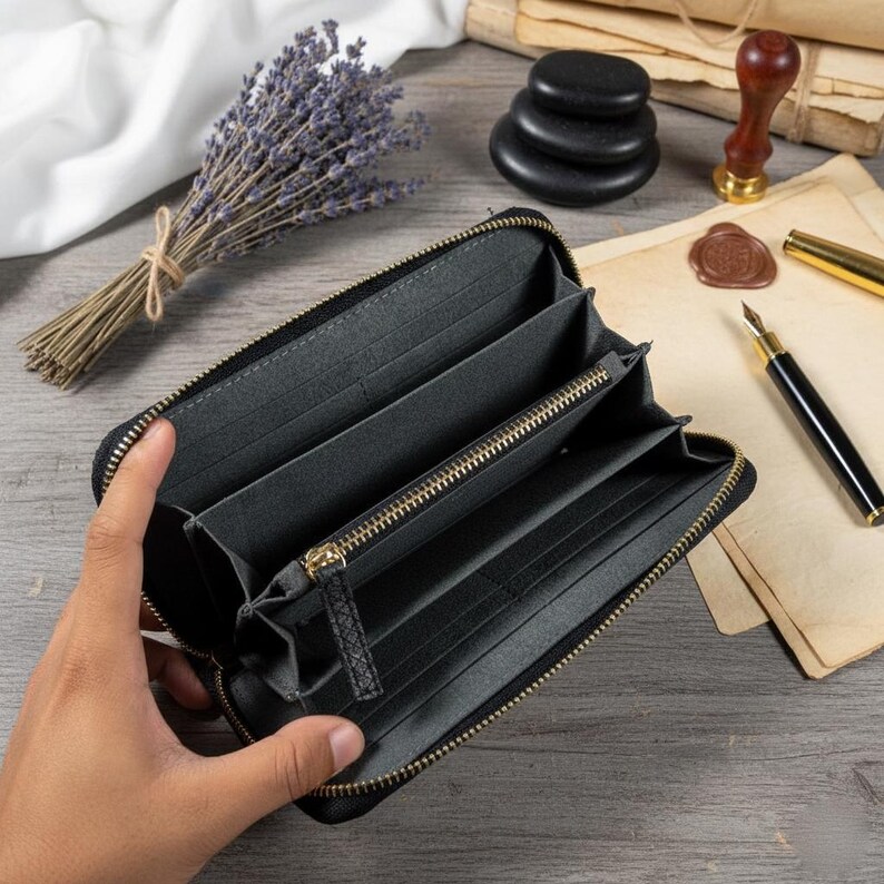 Puede incluir: Cartera negra abierta con m&uacute;ltiples compartimentos y cremallera dorada. La cartera est&aacute; hecha de un material oscuro. El fondo incluye un ramo de lavanda, piedras apiladas y art&iacute;culos de papeler&iacute;a.