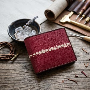 Op de afbeelding: Een bordeauxrode leren portemonnee met een wit stippenontwerp aan de voorkant. De portemonnee is rechthoekig en heeft een bruine rand. Een kleine kom met kristallen en een leren armband liggen ernaast.