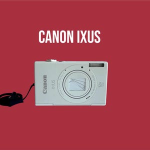 Könnte beinhalten: Eine weiße Canon IXUS Digitalkamera mit einem 12-fach optischen Zoomobjektiv, einem eingebauten Blitz und einer schwarzen Handschlaufe. Das Kameragehäuse zeigt die Canon- und IXUS-Logos, mit dem Text CANON IXUS über dem Gerät.