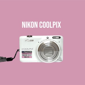 Op de afbeelding: Een witte Nikon Coolpix digitale camera met een 12x optische zoomlens, een zwarte polsband en tekst op de behuizing waaronder 16.0 Megapixels CMOS, Glamour Retouch, Quick Effects en Wi-Fi. De merknaam NIKON COOLPIX staat boven de camera.