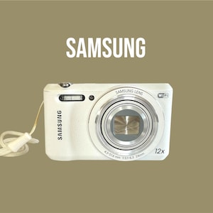 May include: White Samsung digital camera with a silver lens and a cream-coloured wrist strap. The camera has the word "SAMSUNG" printed on the front and the top. The lens has the text "SAMSUNG LENS" and "12x" printed on it.