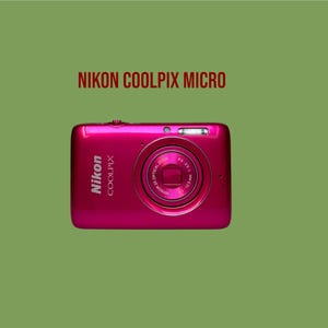 Könnte beinhalten: Eine leuchtend magentafarbene Nikon Coolpix Digitalkamera mit einem mittigen Objektiv und der Aufschrift NIKON 3X OPTICAL ZOOM 4.6-13.8mm 1:3.2-5.8. Das Gehäuse zeigt links das Nikon Coolpix Logo und darüber den Text NIKON COOLPIX MICRO.