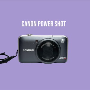 Peut inclure: Un appareil photo numérique Canon PowerShot SX220 HS gris foncé avec un objectif zoom optique 14x. L'avant de l'appareil présente le logo Canon, les spécifications de l'objectif et une dragonne noire. Le texte CANON POWER SHOT est visible.