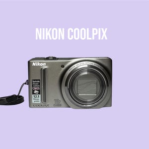 Op de afbeelding: Een grijze Nikon Coolpix digitale camera met een 18x groothoek optische zoomlens, een 12,1 megapixel sensor en een zwarte polsband. Op de voorkant staat de tekst Nikon, Coolpix, 18x Wide Optical Zoom en Backside Illumination CMOS.