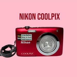 May include: A metallic red Nikon Coolpix digital camera with a black wrist strap. The front features a silver lens ring, a flash, and a sticker stating Slim Body, Li-ion Battery, Wide 5x Zoom, and 14.0 Megapixels. The text NIKON COOLPIX is visible.