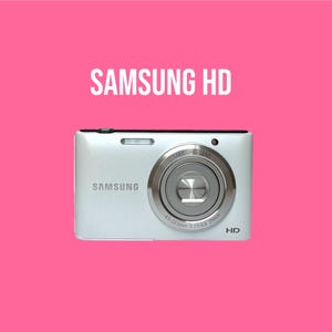Op de afbeelding: Een witte Samsung digitale camera met een zilverkleurige lenscilinder waarop SAMSUNG LENS en 4.5-225mm 1:2.5-6.3 25mm staat. De camerabehuizing toont het Samsung-logo en de tekst HD tegen een effen, felroze achtergrond.