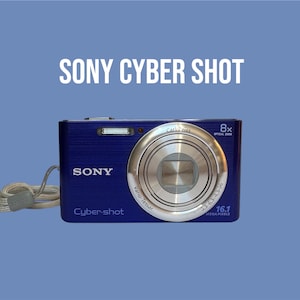 Może przedstawiać: Niebieski aparat cyfrowy Sony Cyber-shot ze srebrnym obiektywem Carl Zeiss, 8-krotnym zoomem optycznym i 16,1 megapikselami. Aparat posiada metaliczne wykończenie, wbudowaną lampę błyskową oraz jasny pasek na nadgarstek przymocowany z boku.