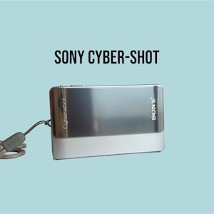 Może przedstawiać: Srebrny aparat cyfrowy Sony Cyber-shot o prostokątnym korpusie, z metalicznym wykończeniem, logo Sony oraz napisem Cyber-shot z przodu. Do boku obudowy aparatu przymocowany jest pasujący srebrny pasek na nadgarstek.