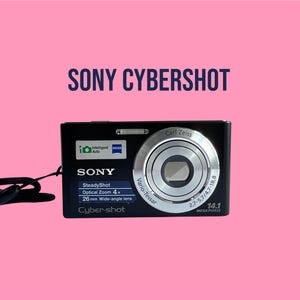 Könnte beinhalten: Eine schwarze Sony Cyber-shot Digitalkamera mit einem silbernen Carl Zeiss Vario-Tessar Objektiv. Auf der Vorderseite befinden sich Aufschriften wie SteadyShot, 4x Optical Zoom, 26mm Wide-angle lens, 14.1 Mega Pixels und ein Intelligent Auto Logo. Eine schwarze Handschlaufe ist befestigt.