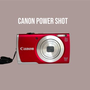 Może przedstawiać: Czerwony aparat cyfrowy Canon Power Shot z 5-krotnym zoomem optycznym, 16,0 megapikselami i czarnym paskiem na nadgarstek. Z przodu aparatu widoczne jest logo Canon, specyfikacja obiektywu oraz napis CANON POWER SHOT nad urządzeniem.
