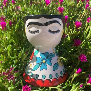 May include: A white ceramic planter shaped like a woman's face with black hair, closed eyes, and a black unibrow. The planter has a colorful floral pattern with blue, yellow, and red accents.