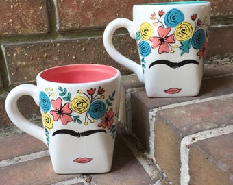 May include: Two white ceramic mugs with a Frida Kahlo inspired design. The mugs feature a colorful floral crown, a unibrow, and a red lip. One mug has a teal blue rim and the other has a coral pink rim.