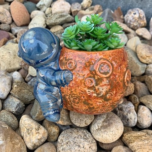 May include: A blue astronaut figurine holding onto a brown and orange ceramic planter shaped like the moon. The planter is filled with green succulent plants.