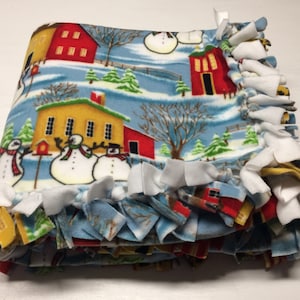 May include: A blue fleece tie blanket with a winter wonderland print featuring snowmen, houses, and trees. The blanket has white, yellow, and blue fringe.