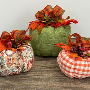 May include: Three decorative fabric pumpkins in fall colors. One is green with a leaf pattern, another is patterned with fall leaves, and the third is orange and white checkered. Each has a decorative bow and berries.