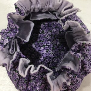 May include: A purple fabric pouch with a white floral print and a light purple lining. The pouch has a gathered top and is made of a soft, quilted fabric.