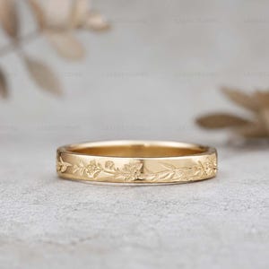 Puede incluir: Un anillo de banda de oro con un patrón floral grabado de forma continua en la superficie exterior. El anillo tiene un acabado pulido y un perfil plano, mostrando tallas detalladas de pequeñas flores y hojas que rodean toda la circunferencia.