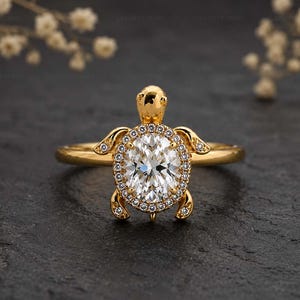 May include: A gold ring featuring a turtle-shaped design with a central oval-cut white gemstone surrounded by a halo of small round white stones. The turtle's head, flippers, and shell are crafted in polished gold, with small stones set into the flippers.