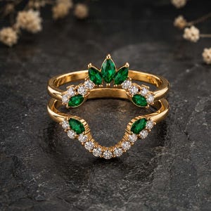 May include: A gold ring guard featuring a curved design adorned with marquise-cut green stones and round white stones. The band is crafted from polished yellow gold, showcasing a symmetrical arrangement of alternating green and white gems.