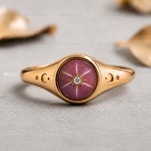 Peut inclure: Une bague en or ornée d'une pierre rose ronde taillée en cabochon avec un effet d'étoile à six branches. Le centre de la pierre est serti d'un petit diamant rond transparent. L'anneau est gravé de petits croissants de lune et d'étoiles.