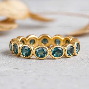 May include: A gold ring featuring a series of oval-cut, teal-colored gemstones. The gemstones are set in individual bezels, creating a continuous band. The ring's gold band provides a warm contrast to the cool tones of the gemstones. The ring is a piece of jewelry.