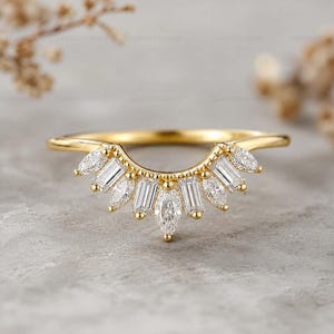 May include: A gold ring with a curved design, set with clear gemstones. The gemstones are a mix of marquise and baguette cuts, arranged in a fan-like pattern. The ring is displayed against a neutral grey background. The ring's band is a thin, elegant design.