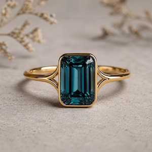 May include: A gold ring with a rectangular, teal gemstone. The ring's band has a split design on either side of the gemstone. The ring is set against a neutral background, highlighting the jewellery's design and colour.
