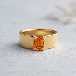 Puede incluir: Un anillo de oro con una gema naranja rectangular en el centro. La banda es ancha y lisa, con un acabado mate. La gema es de un color vibrante y cálido, que contrasta con el oro.