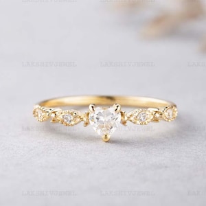 Puede incluir: Un delicado anillo de oro con un diamante en forma de corazón como piedra central. La banda está adornada con pequeñas piedras preciosas claras y detalles decorativos. El anillo se presenta sobre un fondo neutro, resaltando su elegante diseño.