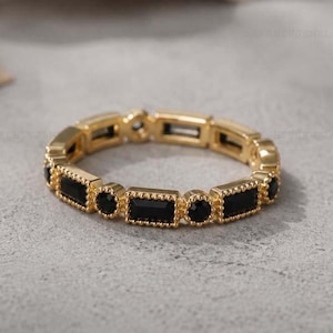 Op de afbeelding: Een goudkleurige ring met een herhalend patroon van zwarte rechthoekige en ronde stenen. De ring heeft een delicaat, sierlijk ontwerp, met een getextureerde band. De ring is geplaatst tegen een neutrale achtergrond.