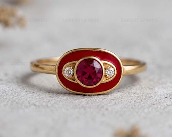Puede incluir: Un anillo de oro con una pieza central ovalada y un esmalte rojo vibrante. El centro contiene una piedra preciosa roja redonda y facetada, flanqueada por dos pequeñas piedras claras de corte redondo, sobre una banda de oro pulido.