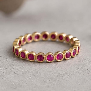 Puede incluir: Una alianza de oro con una hilera continua de piedras preciosas rosas redondas engastadas en bisel. El anillo tiene un acabado metálico pulido y un diseño circular uniforme, mostrando una serie de piedras idénticas fijadas en el metal dorado.