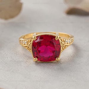 Könnte beinhalten: Ein goldener Ring mit einem großen, quadratisch geschliffenen, leuchtend roten Edelstein. Der Ring hat aufwendige Filigranarbeiten an den Seiten und kleine Akzentsteine. Das Band ist goldfarben poliert und wirkt klassisch und elegant.