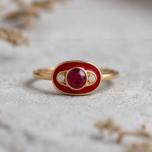 Puede incluir: Un anillo de oro con una piedra central roja de forma ovalada y dos piedras blancas más pequeñas. El anillo tiene un engaste de esmalte rojo y una banda fina. Es una joya.